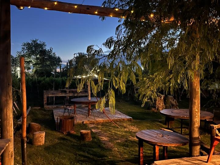 Pérgola de madera con guirnaldas de luces y mesas bajo los árboles al anochecer